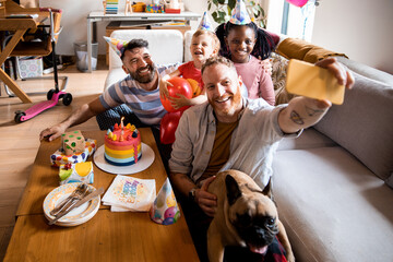 Young male gay couple taking a selfie on a smartphone with their children while celebrating a birthday at home