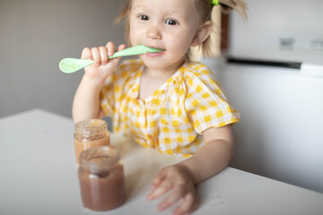 A little girl with a spoon in her hands is sitting at the table eating fruit puree on her own.