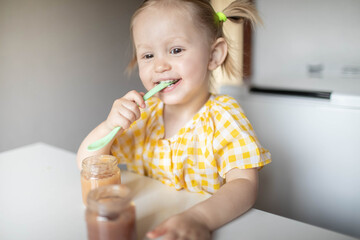 A little girl with a spoon in her hands is sitting at the table eating fruit puree on her own.