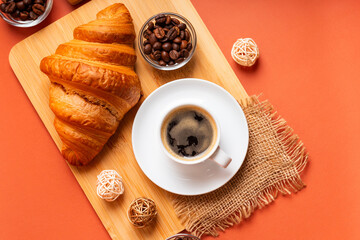 French croissant, cup of black coffee, aromatic roasted arabica and robusta coffee beans in a glass jar on  wooden desk from above on orange background in rustic style. Perfect tasty breakfast.