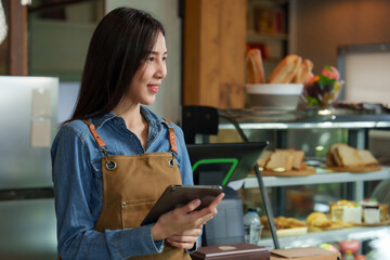 Beautiful female Asian female barista looking and holding  tablet and smiling standing in front of the counter And next to it there is  bakery cabinet with bread, cakes, and products from small cafe