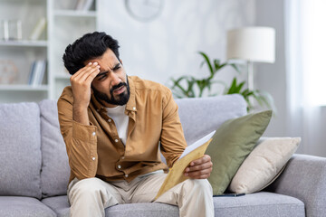 Fototapeta premium Upset and disappointed man sitting on sofa at home, hispanic received mail message letter with bad news, sad depressed in living room at home