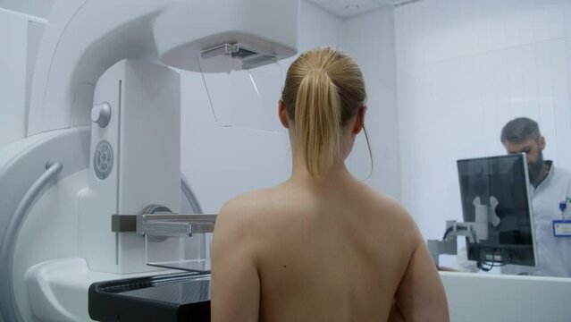 Caucasian adult woman stands topless in hospital radiology room. Female patient undergoing mammography screening procedure in modern clinic. Male doctor adjusts mammogram machine using computer.