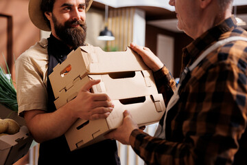 Farmer providing locally sourced bio vegetables to happy vendor selling it to sustainable living clients in zero waste shop. Man handing crates full of fresh natural produce to smiling retailer