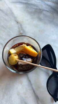 black sunglasses next to a glass of vermouth with olive orange and ice cube on a marble table: catalan vibes, barcelona