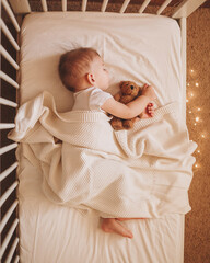 A little baby in a white bodysuit sleeping in a white crib holding a teddy bear and surrounded by...