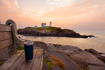 A bench and a thermos of coffee with a view of the sunrise on the coast and a lighthouse in the background. USA. Maine. Nubble Lighthouse