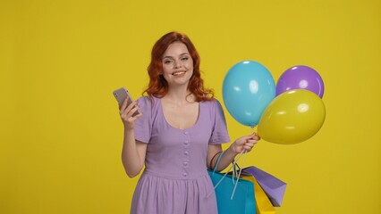 Woman with shopping bags and balloons in studio on yellow background. A woman uses a smartphone.