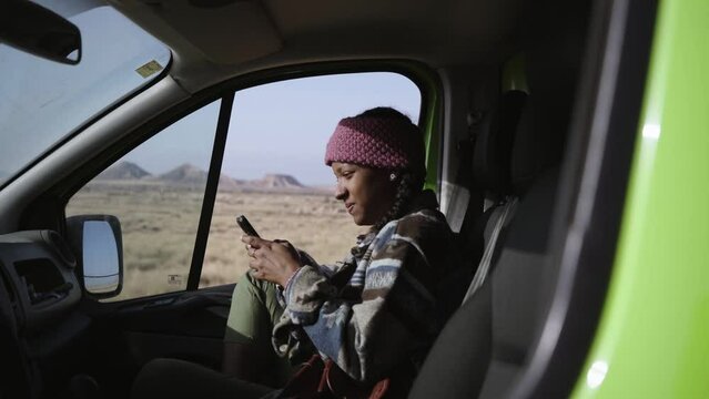 Young African woman using cell phone smiling inside camper. Black girl co-driver typing mobile laughing while resting with background of bardenas reales. Generation z road trips on nature vacation.