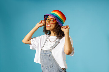 Smiling woman in eyeglasses puts rainbow cap while looks camera on blue studio background