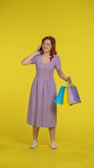 A woman with shopping bags speaks on a smartphone, shares her emotions from shopping. Full length woman in the studio on a yellow background. Vertical shot. Sale, Black Friday, advertising, discounts.
