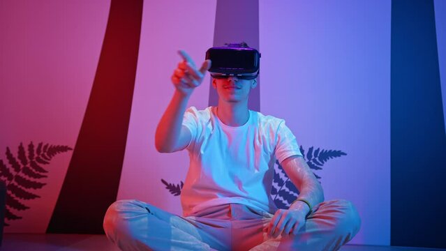 Smiling teenage programmer boy sitting on the floor with a virtual reality headset on, making hand gestures, typing, programming and scrolling in the air, low angle shot.