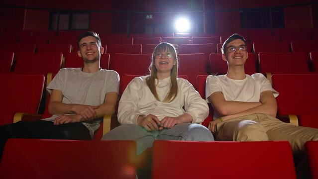 Three friends in the cinema hall, a girl and two boys having fun watching a film in the empty movie place, front view. Entertainment and people concept.