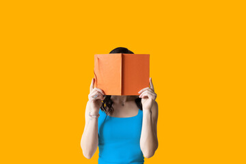 a woman with dark hair covers her face with an orange book on a yellow background