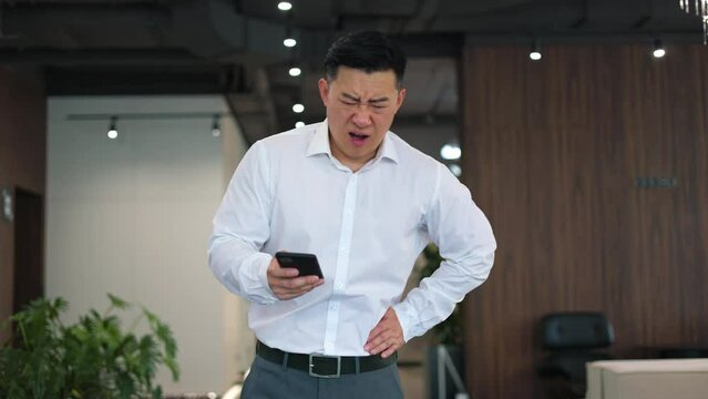 Unhealthy asian employer complaining on abrupt colicky pain while browsing internet on smartphone in office. Young man frowning while feeling sudden and severe spasms in left lower part of abdomen.