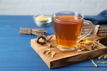 Aromatic licorice tea in cup and dried sticks of licorice root on blue wooden table, space for text