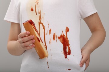 Woman holding hotdog and showing stain from sauce on her shirt against light grey background, closeup