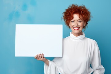 Vibrant portrait of a cheerful mature redhead woman in her 60s, showcasing an ad board with copy space. Her lively demeanor makes the bright blue backdrop pop, perfect for promotions and announcements