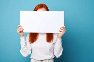 Portrait of a radiant young redhead woman holding an ad board, invitingly showcasing copy space against a bright blue backdrop. Perfect for captivating promotions and lively announcements.