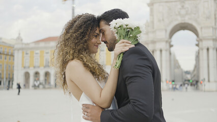 Fototapeta premium Beautiful couple of lovers tenderly embrace on street. Action. Elegant couple in beautiful outfits are gently embracing. Couple in love in beautiful outfits hugs on city square