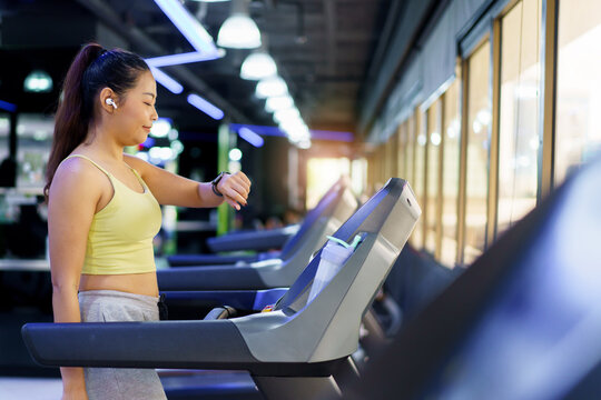 Happy beautiful Asian woman doing an cardio exercise by running on treadmill at the indoor gym, woman using a smartwatch or tracking device while walking and running on treadmill.