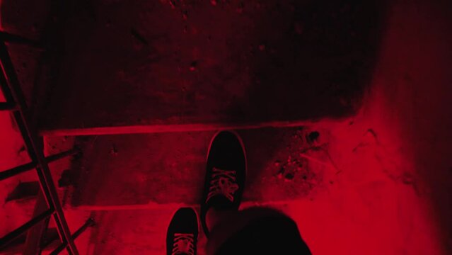 Menes feet climb a narrow concrete staircase in an abandoned building. POV. The steps are covered in dust, pieces of plaster and garbage. An eerie dim red light. Feet in sneakers are walking stairs
