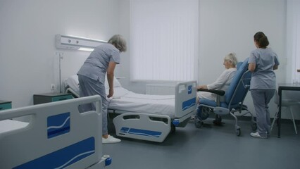 Mature nurse sets up bed using remote control, prepares hospital room for patient. Female medical worker pushes wheelchair with elderly woman and puts her in bed. Modern clinic or medical facility.