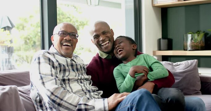 Happy african american father, son and grandfather sitting on couch together laughing, slow motion