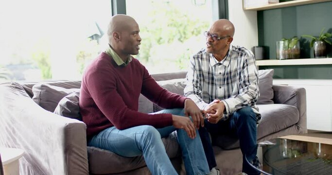 Happy african american senior father and son sitting on couch,talking and laughing, slow motion