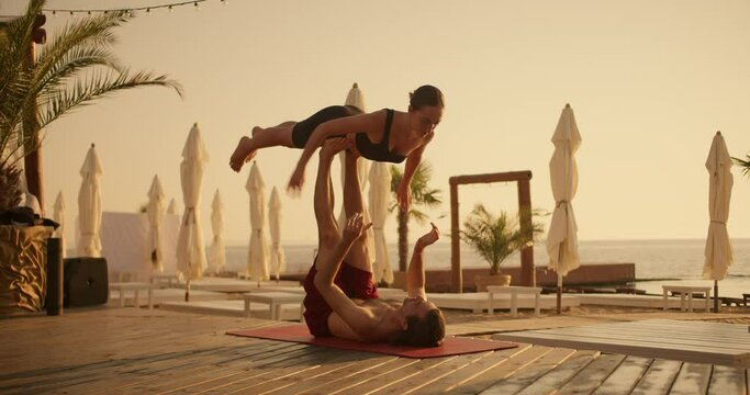 The guy holds the girl up with his legs, the couple does yoga combined with acrobatics on a red carpet on a sunny beach in the summer. Acroyoga and mind relaxation. Yoga flying pose