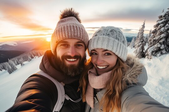 Smiling young couple wearing winter clothes taking selfie photo on snowy mountain at sunset