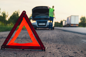 Unrecognizable sad driver in reflective vest. Male driver standing near broken car with open up hood. Red triangle to warn other road users of car breakdown or engine failure stop at countryside