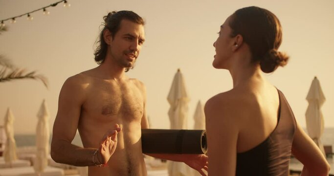 A brunette guy with a naked torso holds a yoga mat in his hands and talks to a brunette girl in a black top on a sunny beach with palm trees and umbrellas in the summer