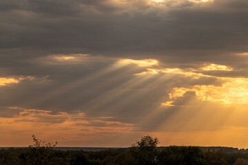 A serene landscape bathed in the warm, radiant glow of a sunset. The suns rays pierce through the cloud-filled sky, casting a beautiful orange hue over the fields and trees below.