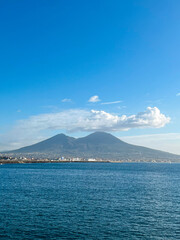 Fototapeta premium View at the mount Vesuvius from the opposite coast in Naples, Italy. Bright blue sky and fluffy clouds located around the volcano crater. The sea is calm. Small fishermen boats are at standstill