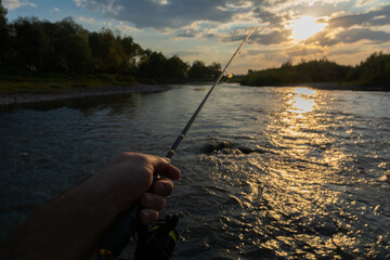 Fishing on river and an amazing sunset with the sun shining through the clouds. Hand holding fishing rod.