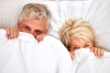 Hiding, portrait and a senior couple in bed for comfort, sleep or love in retirement. Shy, intimate and an elderly man and woman in the bedroom of a house with a blanket covering head in marriage