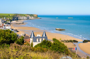 View over Arromanches beach and Cap Manvieux in Normandy, France, on a sunny day, with remains of the WWII artificial Mulberry harbor lying on the sand and in the sea.