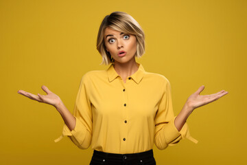 Indecisive Beauty: Clueless Blonde Woman with Short Dark Hair Shrugs Shoulders, Expressing Hesitation Against a Vibrant Yellow Background. - obrazy, fototapety, plakaty