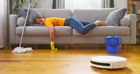 Tired woman lies on sofa while a robot vacuum cleaner does the cleaning