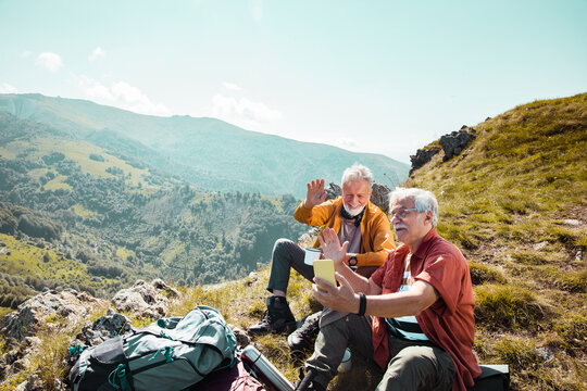 Two senior male friends taking a break from hiking to have a video call on a smartphone in the mountains