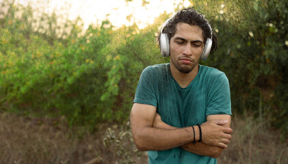Sad young Arabic man looking at camera wearing headphones during the rain in the park. Unhappy male feels cold during the rain outdoors. Guy hates the rainy weather.