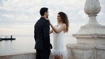 Portrait of happy man in suit and woman in white short dress and curly flying hair. Action. Lovely couple standing on a pier with the calm sea and cloudy sky on the background.