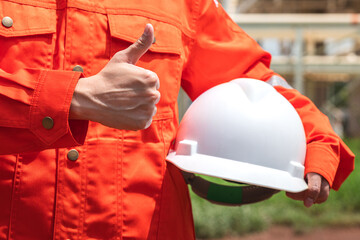 Action of the engineer in orange coverall uniform is holding a white safety helmet with background of chemical or petrochemical refinery plant. Ready to work in challenge job action concept.