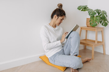 Happy asian Thai woman bun hair, sitting on floor leaning on wall, using tablet, working or watching series at apartment in winter day.