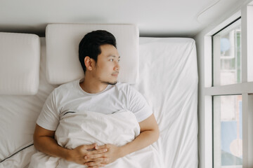 Asian man lying on white bed at apartment, worried and thinking something or missing someone, looking at the window alone.
