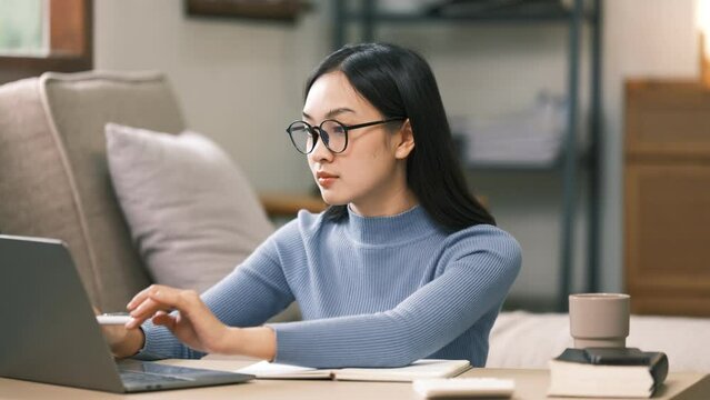 Young adult asian student woman taking notes while using laptop computer at home. Asian female learning online listening virtual video call. Business and education concept.
