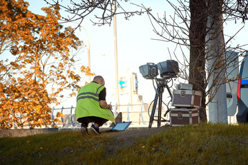 Man sets mobile speed camera next to tree near highway. Police radar installed on roadside to control speed limit. Man adjusts speed radar with laptop. Automatic radar, radar trap for speed monitoring
