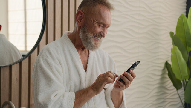 Caucasian relaxed senior old man in bathroom in bathrobe calm mature retired male in bathroom browsing mobile phone scrolling smartphone in morning check social media morning using cellphone smiling - Powered by Adobe