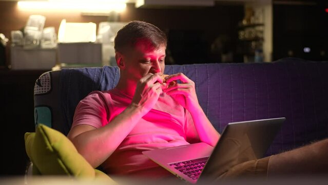Happy hungry young man watching online tv show on laptop, having tasty fresh burger lying on sofa in evening in dark living room. Handsome male watch movie, eat fast food and relax with online video.